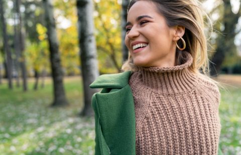 A person wearing a brown knit sweater and green coat stands outdoors, smiling, with blurred trees and grass in the background.